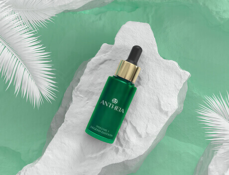 A green bottle with a dropper cap labeled "ANTHEIA" rests on a textured white surface, surrounded by white palm-like leaves on a green background—an enchanting capture crafted by Toronto Design Agency. Conte Studios